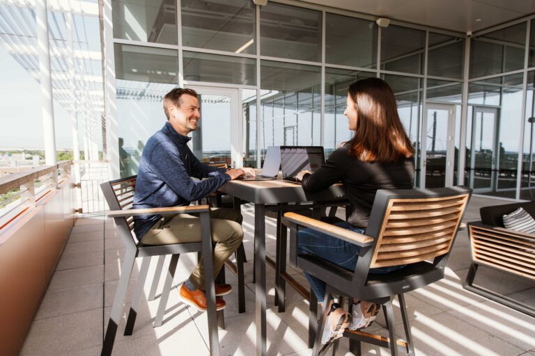 Man and woman smiling while collaborating on laptops on an outdoor rooftop workspace.