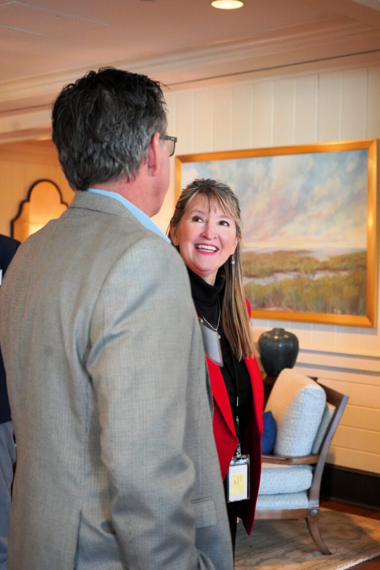 Woman in a red blazer smiling during a conversation with a man at an indoor event.