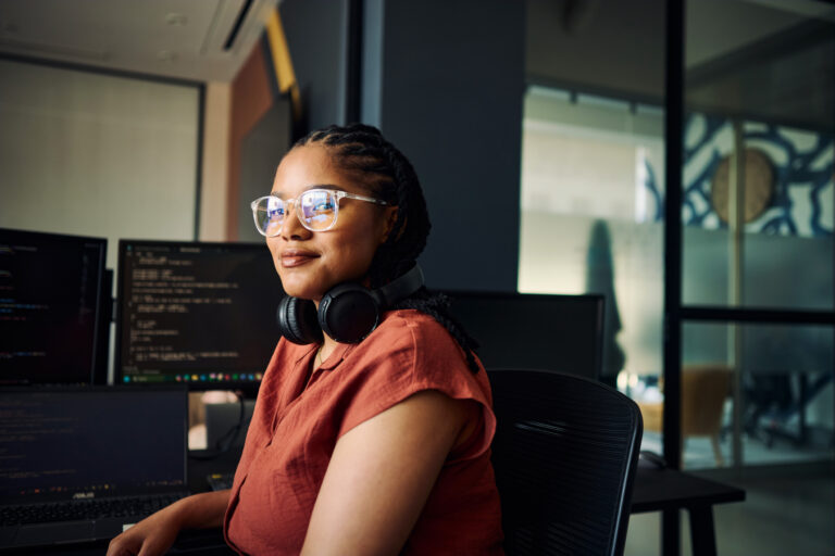 young BIPOC woman sits in front of computers, looking at camera