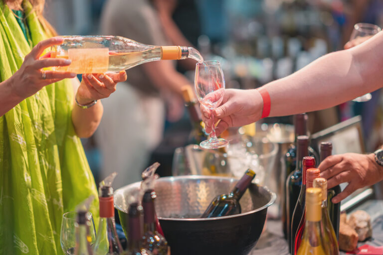 Wine pouring into glass at tasting counter with bartender serving wine to customer surrounded by multiple wine bottles in ice bucket. Wine tasting, hospitality business, alcohol service