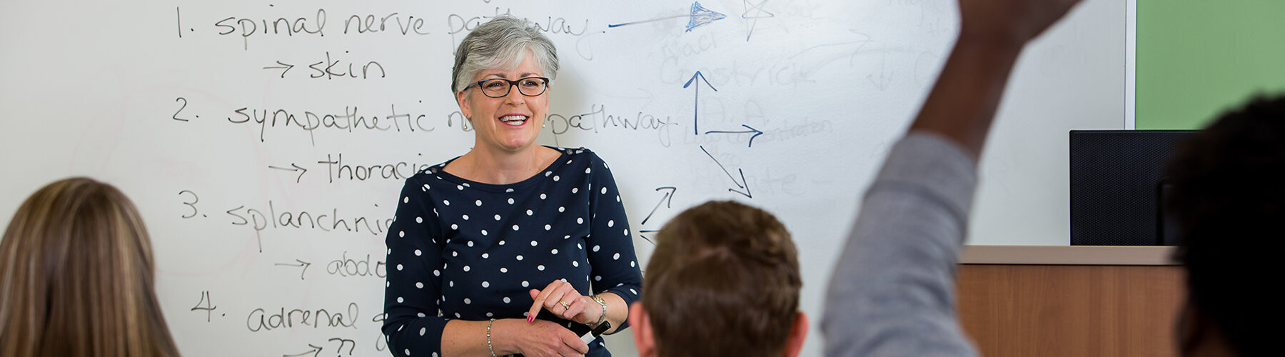 A science teacher in front of a whiteboard in a classroom sees a student raise his hand.