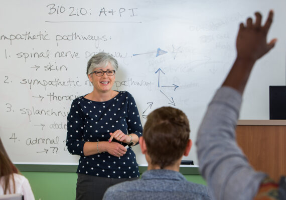 A science teacher in front of a whiteboard in a classroom sees a student raise his hand.