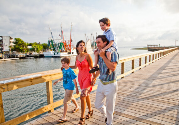 A family of four smiles as they walk down a boardwalk during the day.