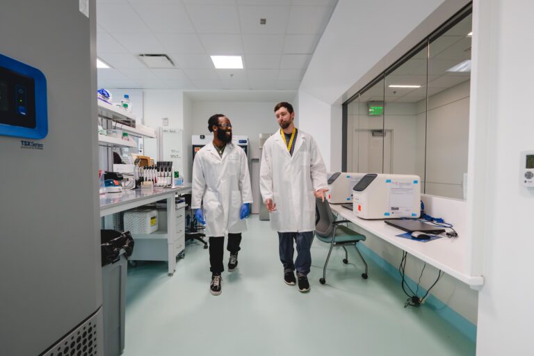 Two young men in lab coats walk through a lab together.