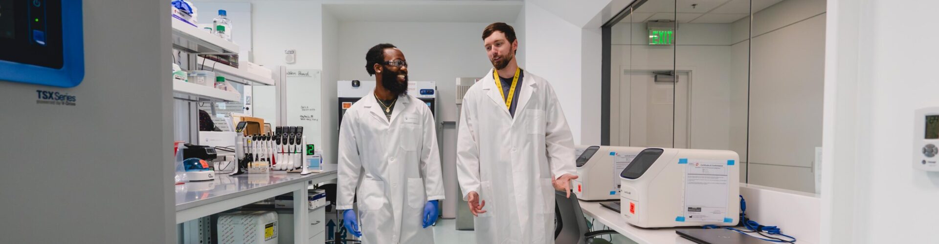 Two young men in lab coats walk through a lab together.