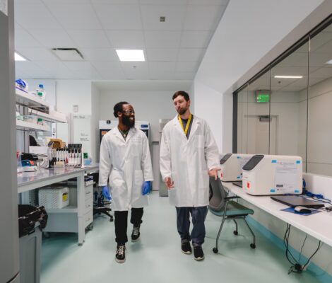 Two young men in lab coats walk through a lab together.