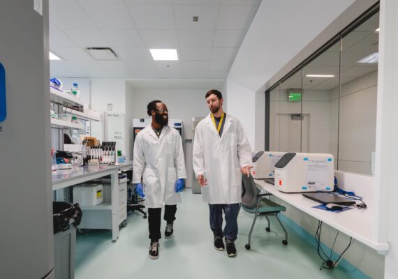 Two young men in lab coats walk through a lab together.