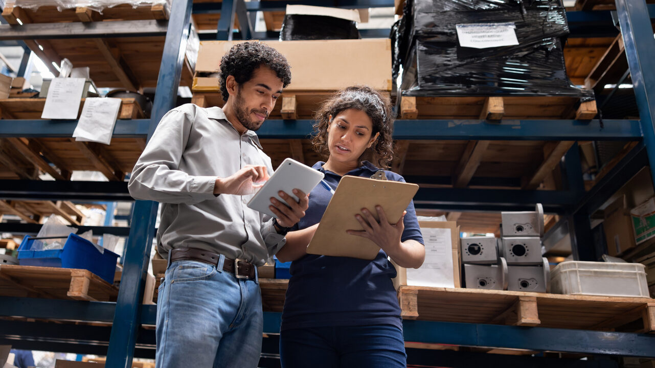 Two coworkers review an iPad and a clipboard together in a fulfillment warehouse.