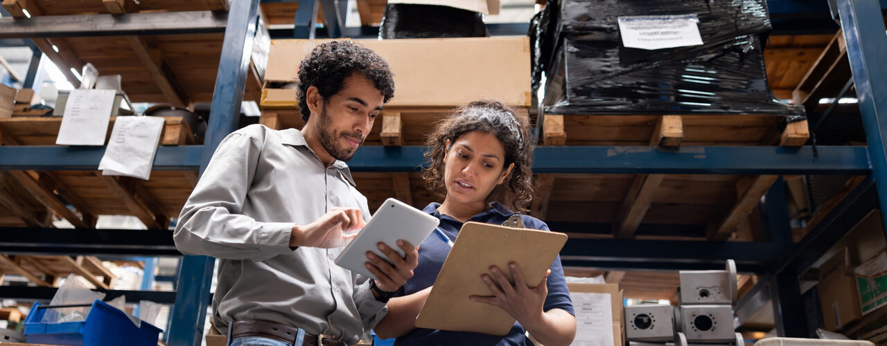Two coworkers review an iPad and a clipboard together in a fulfillment warehouse.