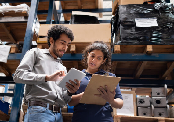 Two coworkers review an iPad and a clipboard together in a fulfillment warehouse.
