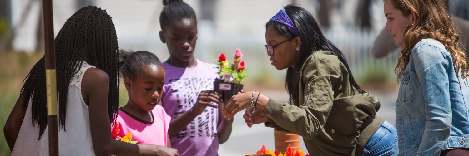 A group of women and children look at flowers at a farmer's market.