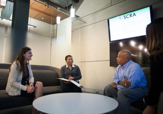 Three business people sit around a casual office table and talk together.