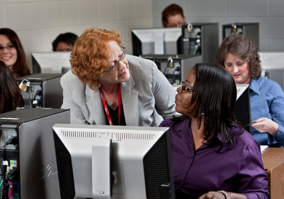 A woman at a desktop computer receives guidance from a supervisor.