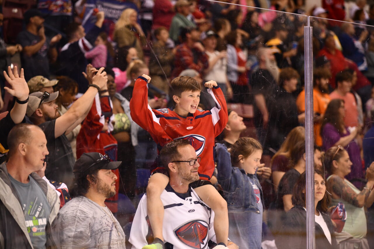 A child in his team's jersey sits on the shoulders of his father and cheers during a hockey match.