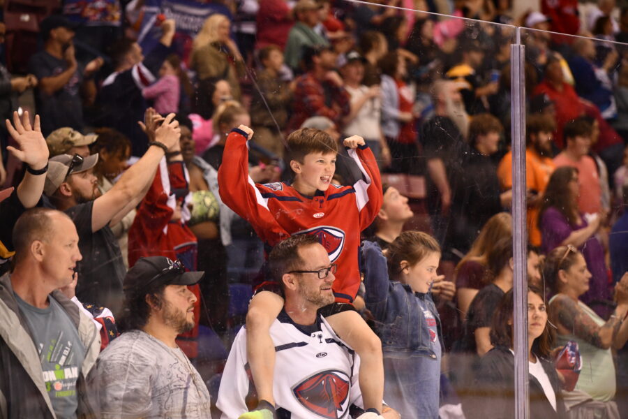 A child in his team's jersey sits on the shoulders of his father and cheers during a hockey match.