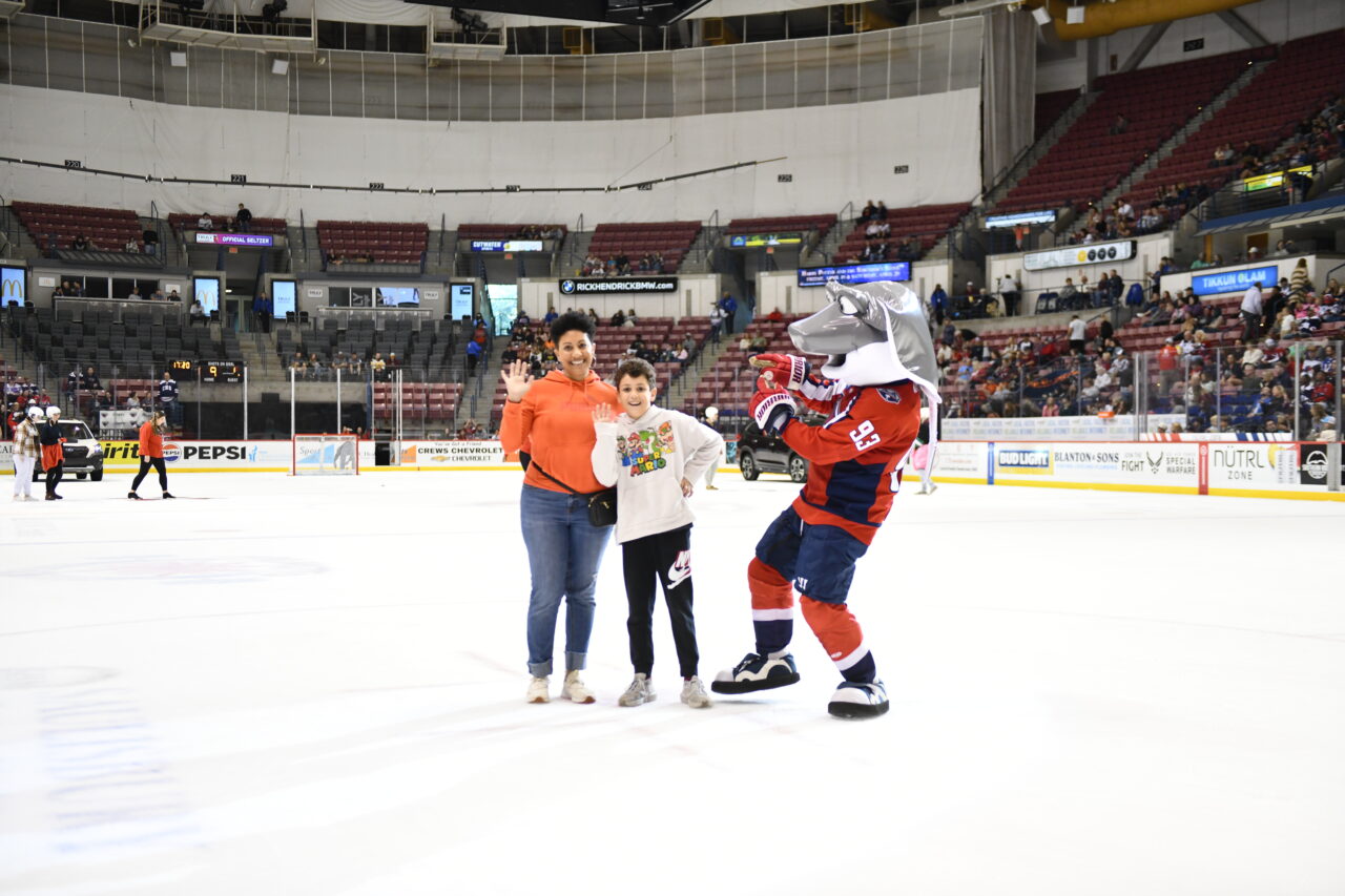 A child and his mom wave at the camera while posing with a mascot for their hockey team.