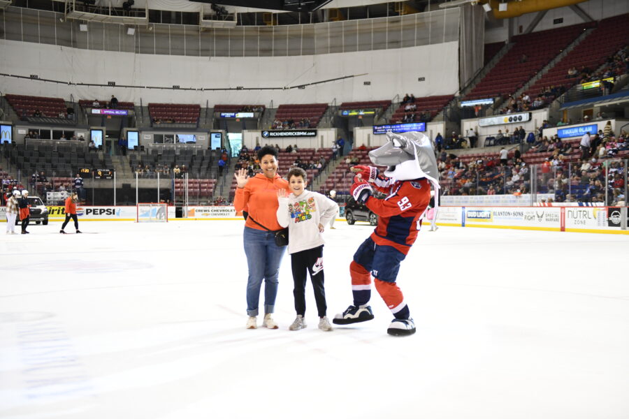 A child and his mom wave at the camera while posing with a mascot for their hockey team.