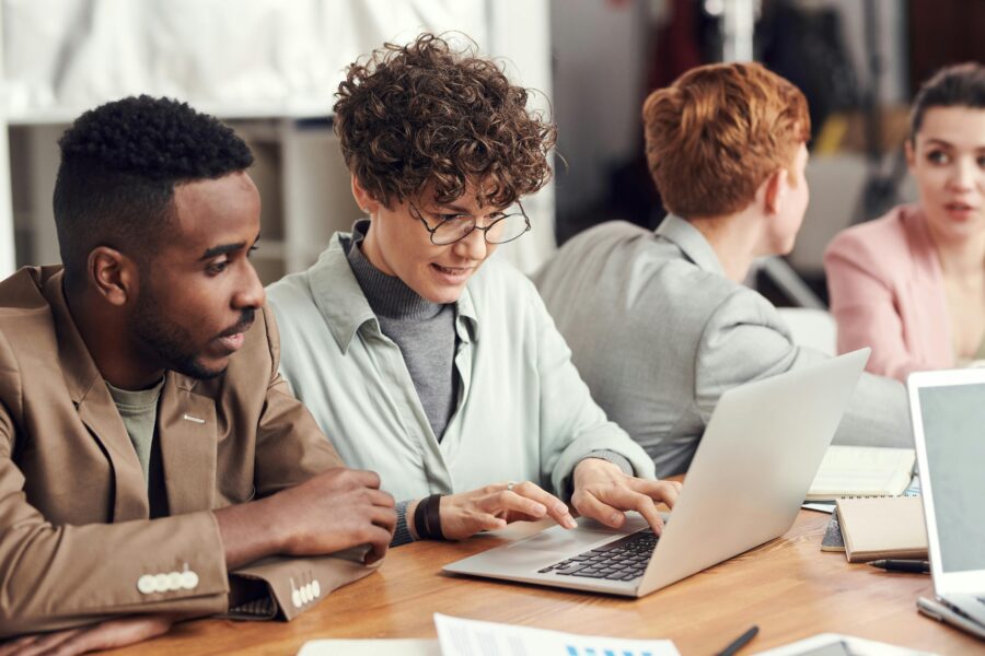 Two coworkers sit at an office table and consult a laptop together.
