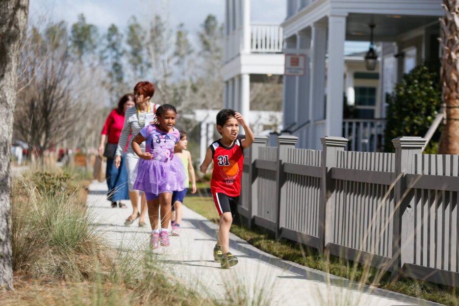 Children pass by houses while running along a sidewalk in a suburban neighborhood.