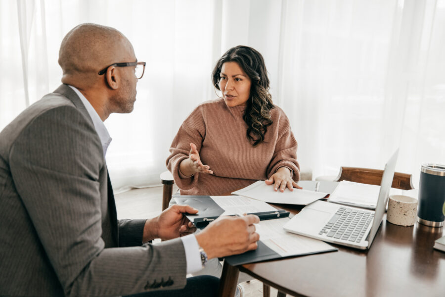 A woman speaks to a businessman at an office table filled with papers and a laptop.
