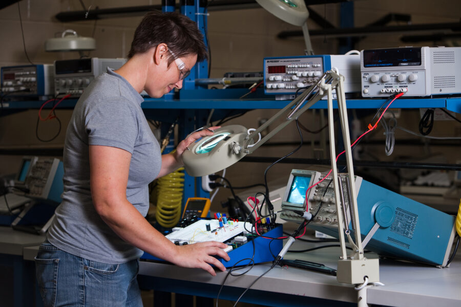 A young woman uses a magnifying glass to work on a circuit board.