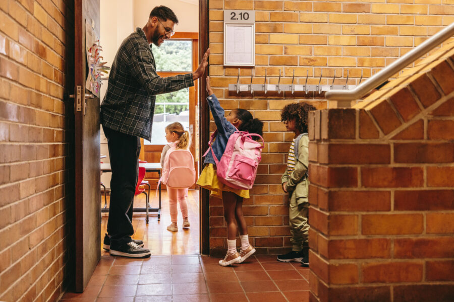 An elementary school teacher greets his students at the door of his classroom with a high five.
