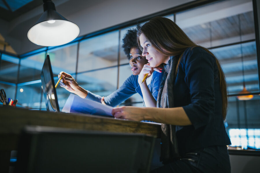 Two young women look closely at a laptop at an office desk together.