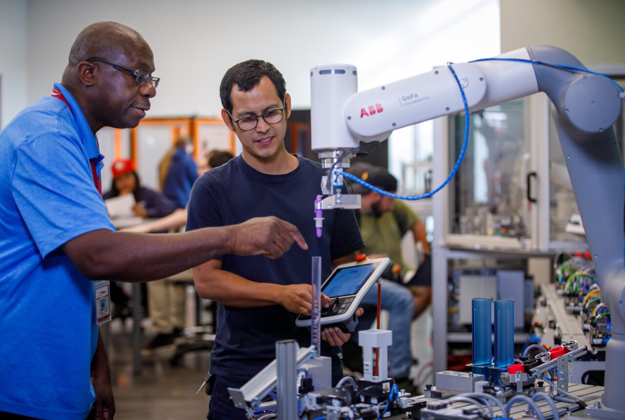 An instructor gives directions to a young man operating a robotic arm.