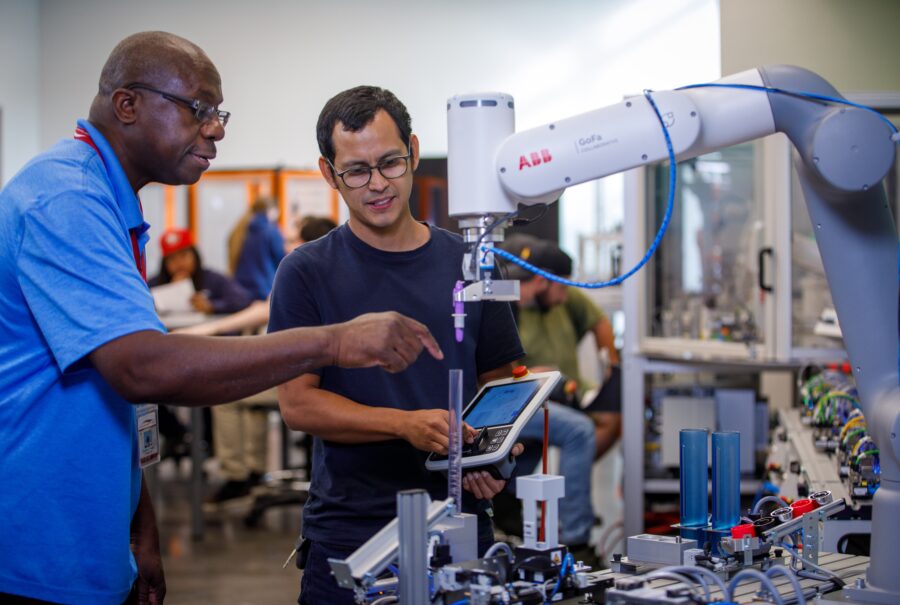 An instructor gives directions to a young man operating a robotic arm.
