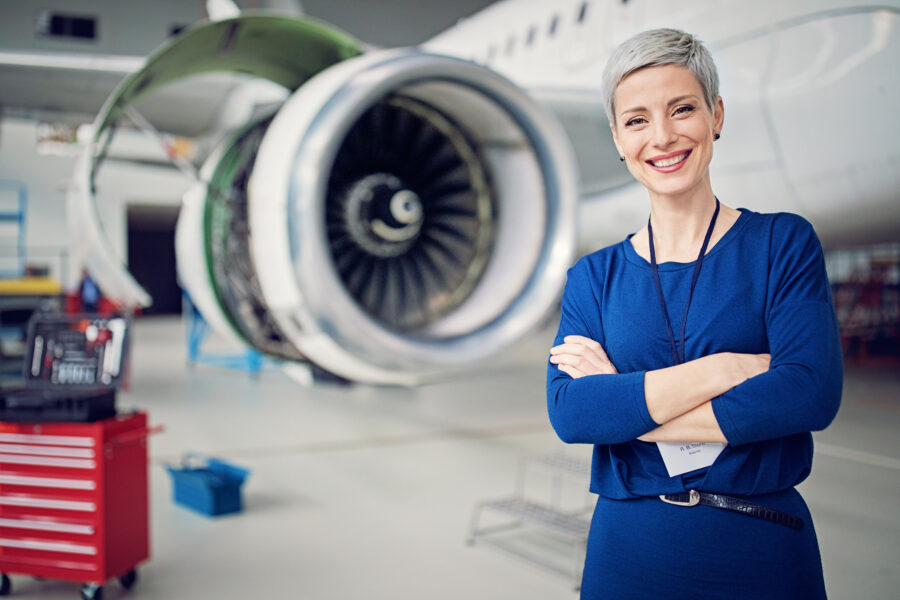 A business woman smiles at the camera while standing in front of a large jet plane on a hangar.