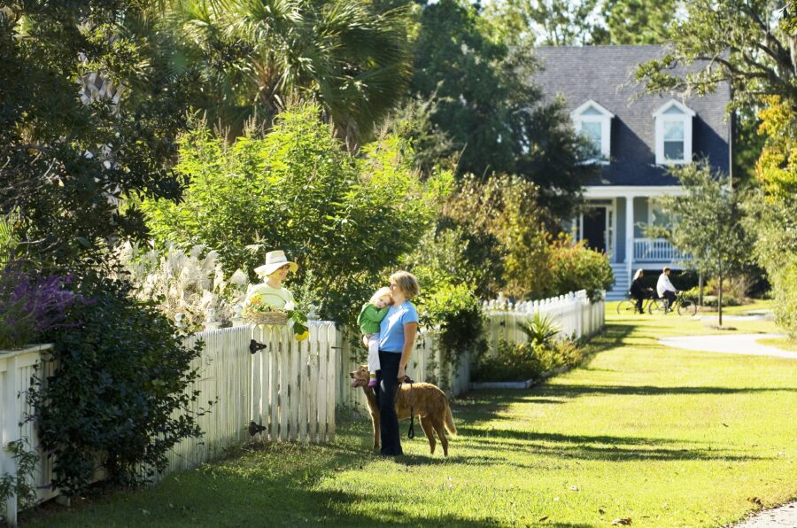 A woman with a dog and child stands and talks to a neighbor at her white picket fence.